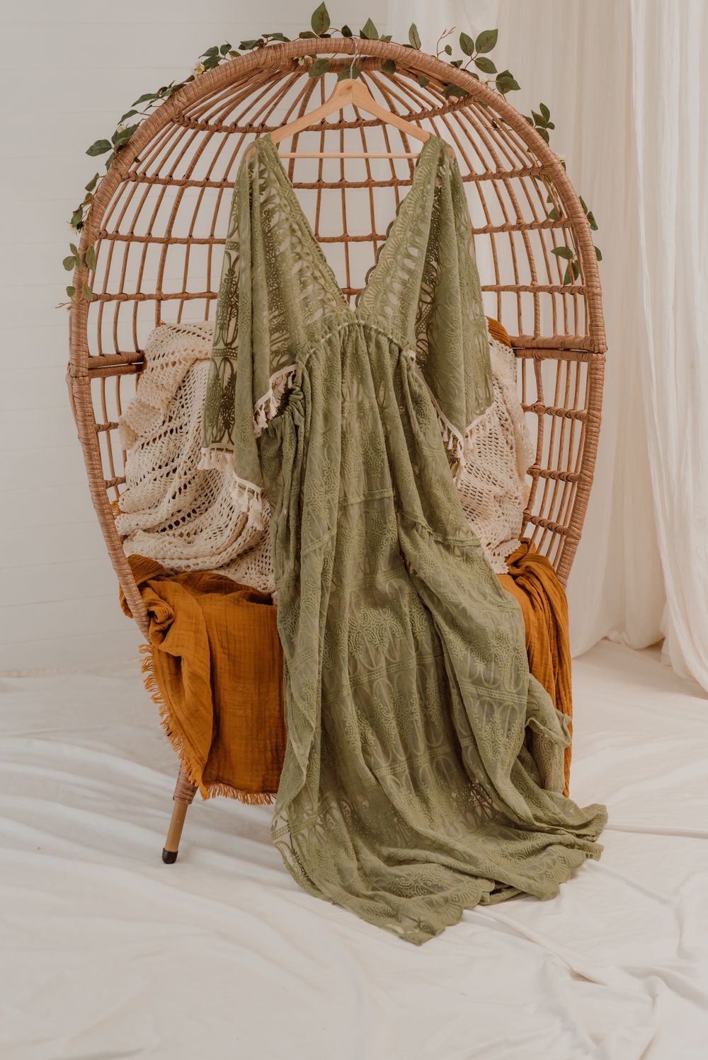 Sage green maxi dress with crochet details displayed on rattan peacock chair with vine decorations.
