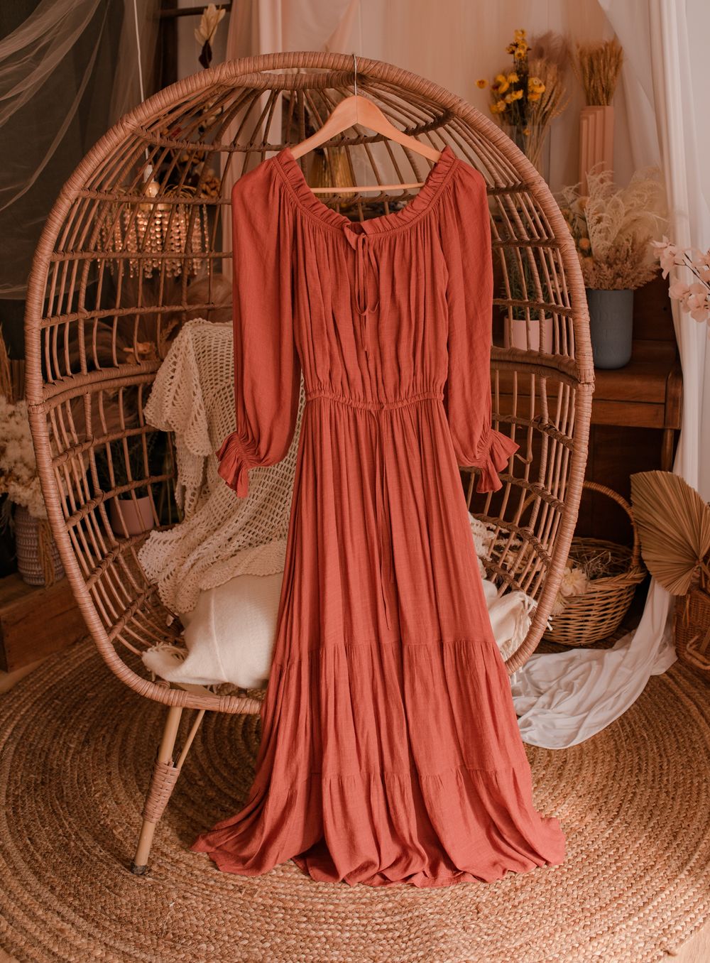 Terracotta-colored flowy maxi dress with gathered waist displayed on rattan peacock chair.