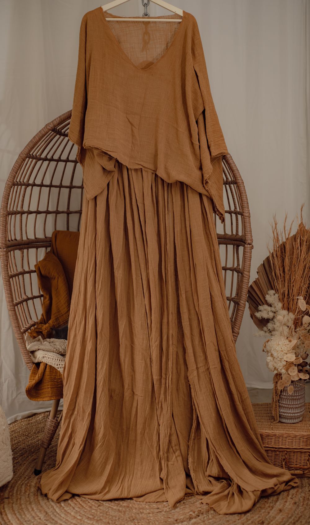 Flowing caramel-colored maxi dress with loose layers hanging on display next to a rattan egg chair and dried flowers.