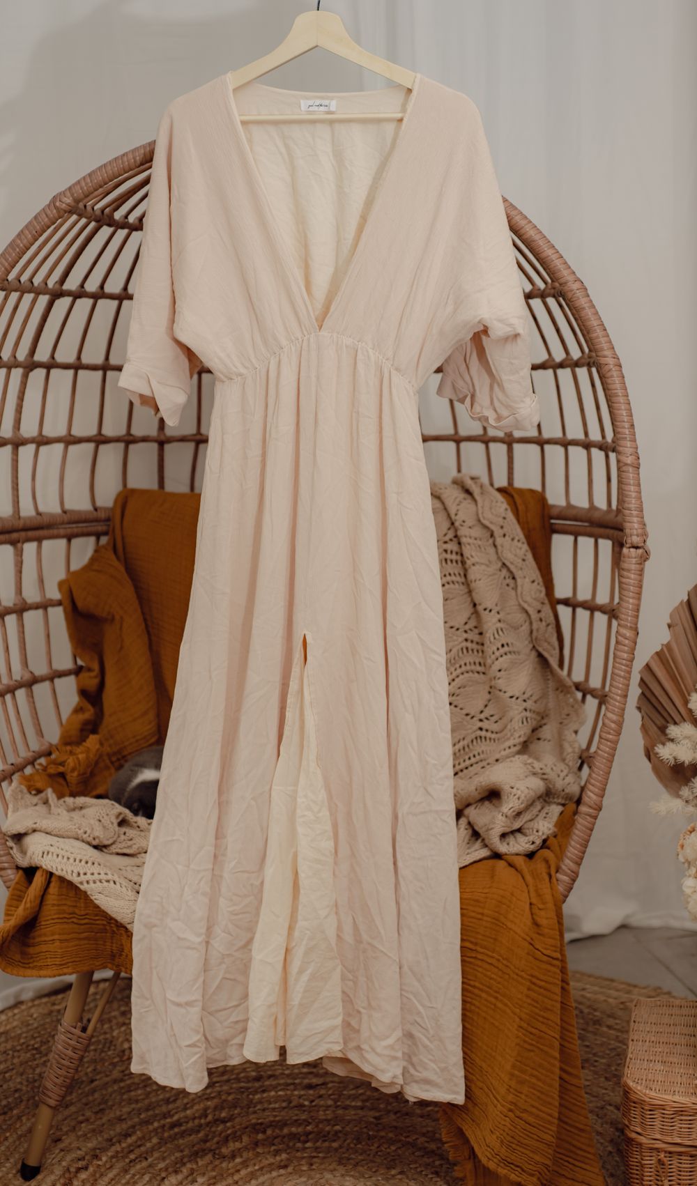 Cream colored flowy maxi dress with empire waist displayed on hanger beside rattan chair and textured decor.