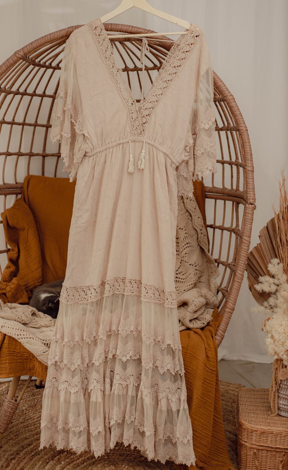 Romantic cream-colored lace maxi dress with ruffled tiers and delicate crochet details displayed on rattan peacock chair.