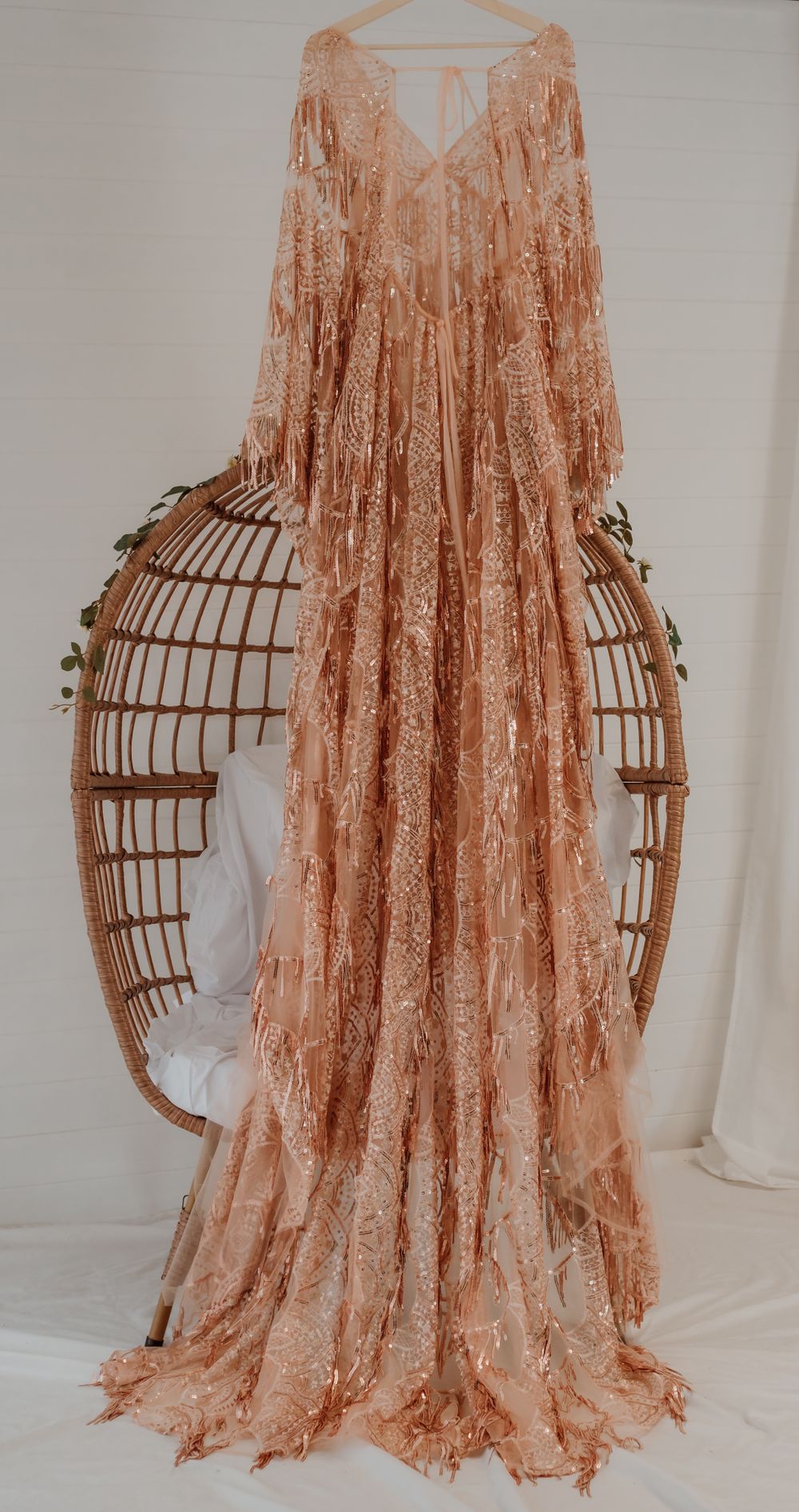 Elegant rose gold lace gown with flowing sleeves draped over a vintage rattan peacock chair.