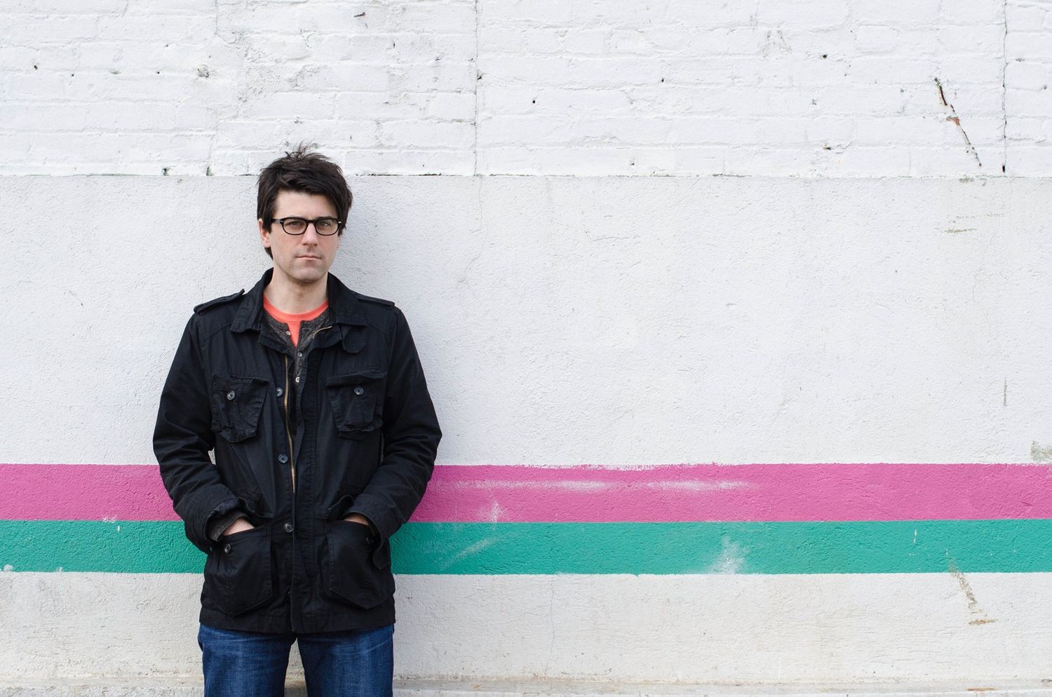 Man in black jacket leans against wall with pink and green stripes against white concrete surface in Jersey City, New Jersey.
