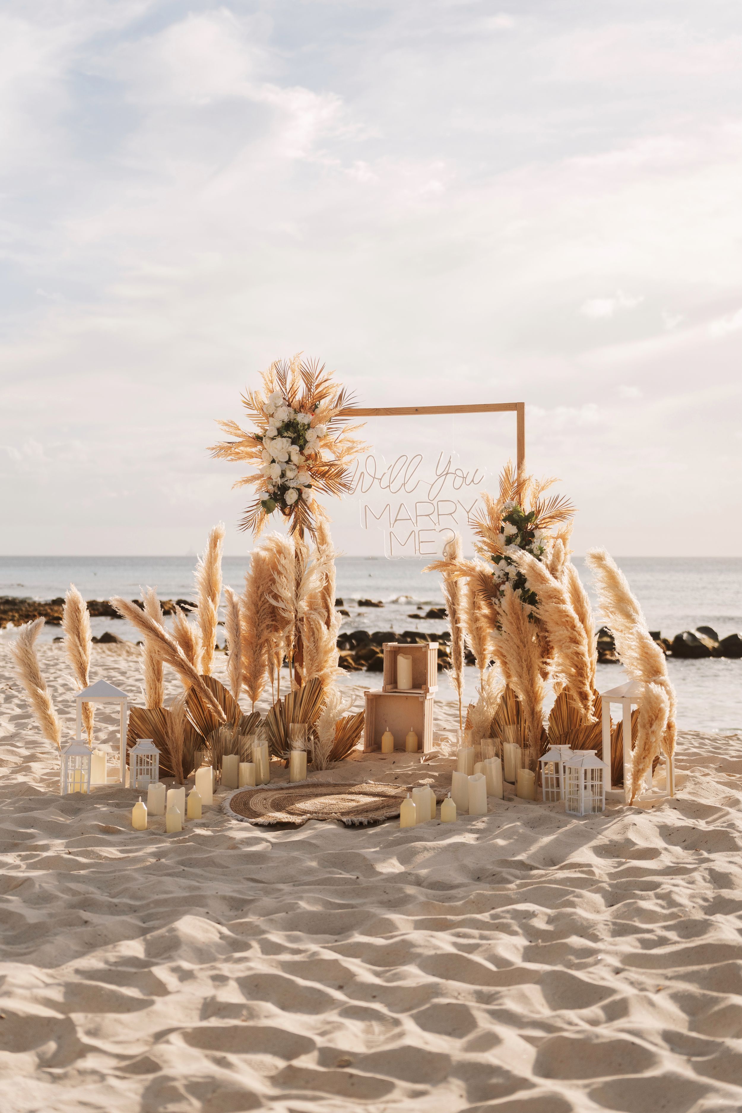Beach proposal setup with pampas grass, candles, and a neon Will You Marry Me sign on white sand by the ocean.