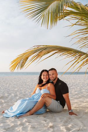 Happy couple sitting together on white sand beach under palm tree fronds, woman in blue dress, tropical ocean backdrop.