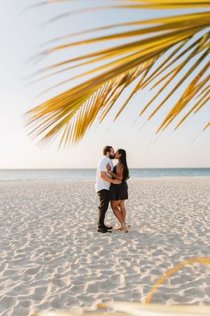 Couple kissing on a white sand beach under a palm frond at sunset with calm ocean in background.