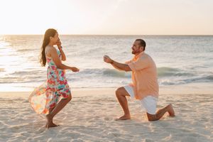Man proposing to woman on a sandy beach at sunset, kneeling with a ring box while she reacts with joy.