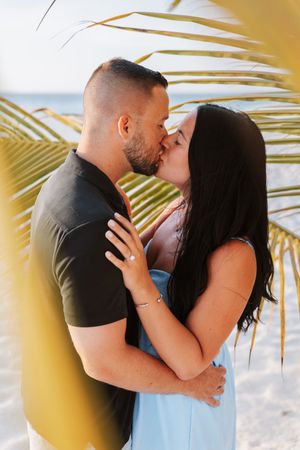 Couple sharing a romantic kiss on a tropical beach framed by palm leaves, woman wearing a light blue dress.