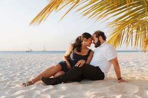 Romantic couple sitting on white sand beach under palm tree at sunset with sailboat in background.