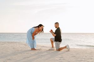 Man proposing to woman on a sandy beach at sunset, kneeling with a ring box as she leans in with joy.