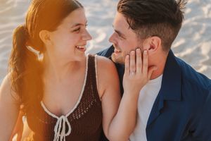 A smiling couple shares a romantic moment on a sunny beach, the woman gently touching the man's face.