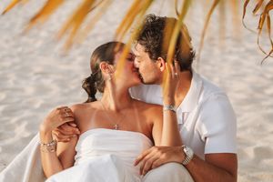 Romantic couple sharing a tender kiss on a tropical beach, dressed in white, surrounded by palm leaves and sandy shores.