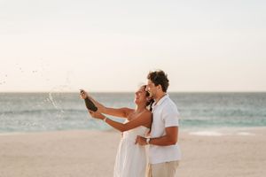 Couple celebrating on beach, popping champagne bottle at sunset, wearing white outfits with ocean in background.