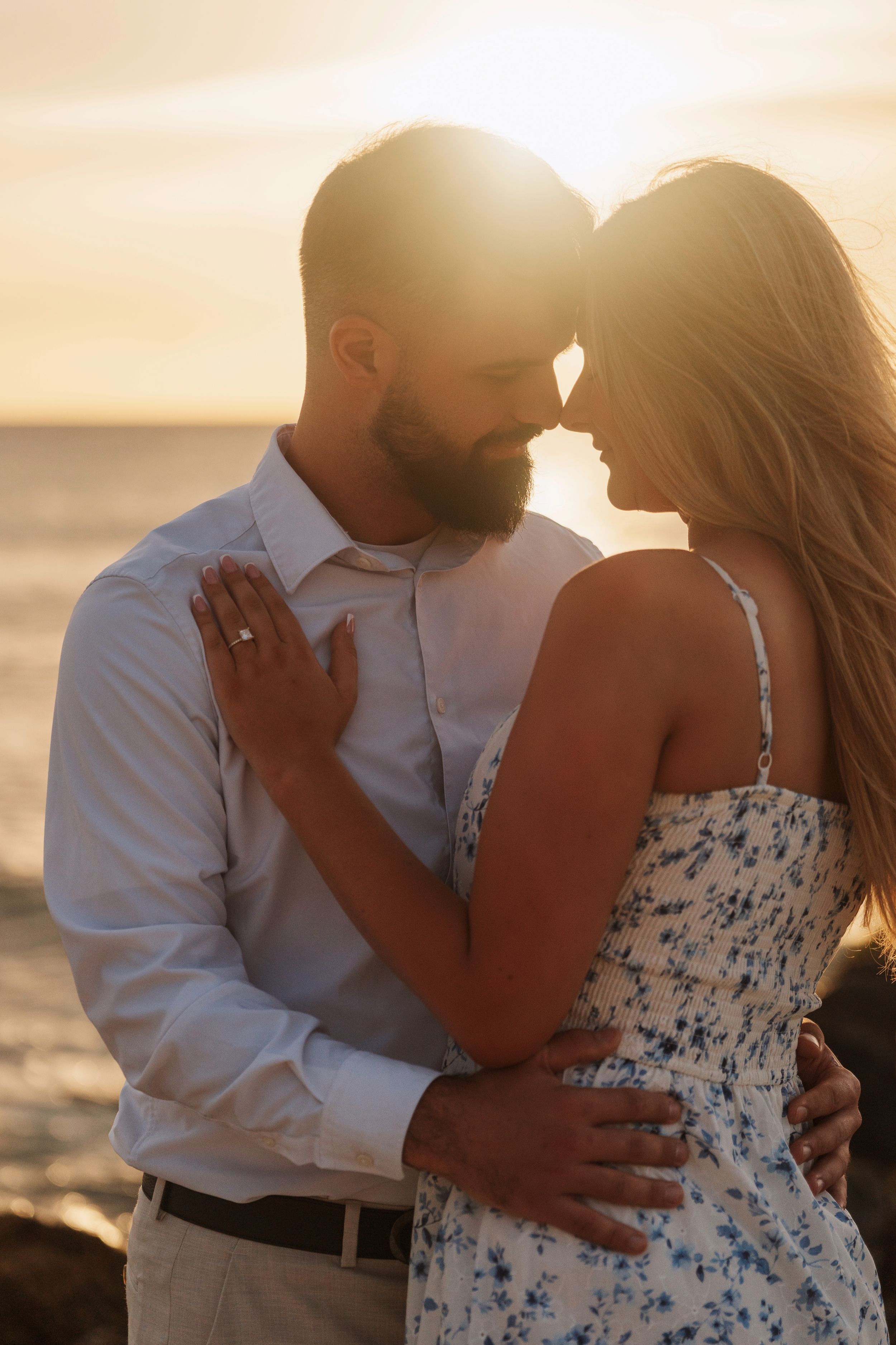 Couple embracing at golden sunset by the ocean, woman wearing engagement ring in floral dress.