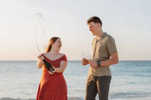 Couple celebrating on beach, woman in red dress popping champagne bottle, man holding glasses by ocean.