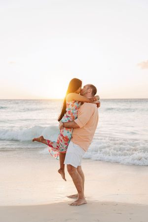 Couple sharing a romantic kiss on a tropical beach at sunset, man lifting woman in floral dress by ocean waves.