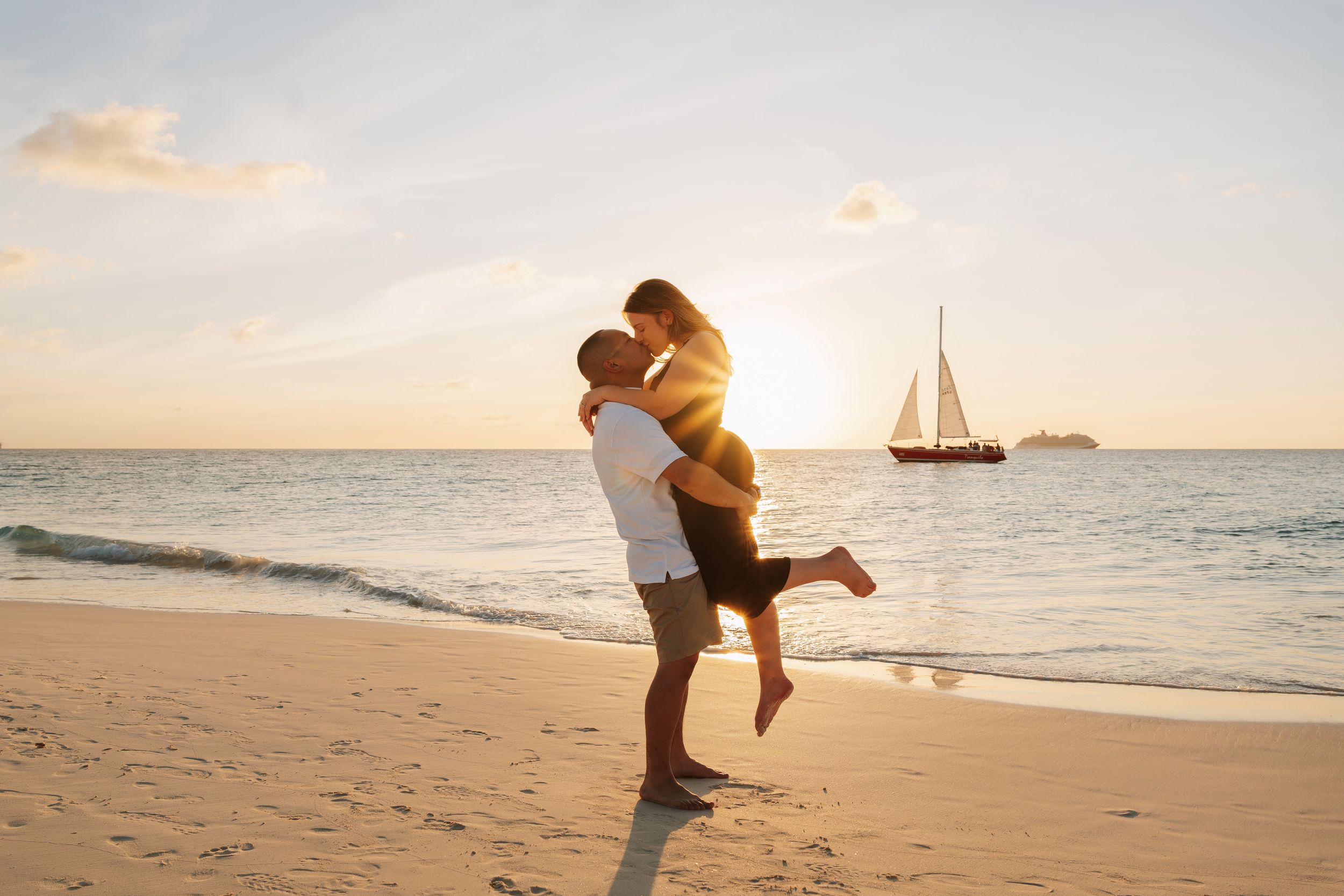 Couple embracing on a tropical beach at sunset with a sailboat in the background.