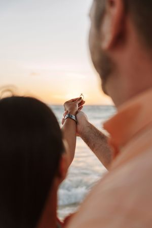 Couple holds hands at sunset beach, woman displaying engagement ring over ocean waves.