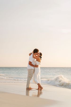 Romantic couple kissing on a sandy beach at sunset, wearing white outfits with ocean waves in the background.