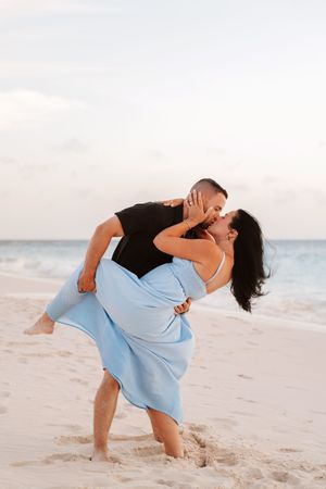 Romantic couple kissing on a sandy beach at sunset, man lifting woman in a flowing blue dress by the ocean.