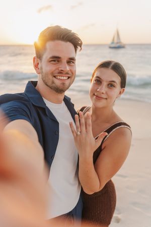 Happy couple takes beach selfie at sunset, woman showing off engagement ring with sailboat in background.