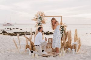 Man proposes to woman on beach with floral arch, pampas grass, and candles at sunset.