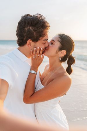A couple shares a romantic kiss on a beach at sunset, both dressed in white, with an engagement ring visible.