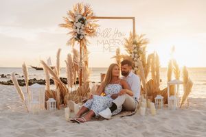 Couple sitting on beach sand at sunset beside a boho-style proposal setup with pampas grass and a Will You Marry Me sign.