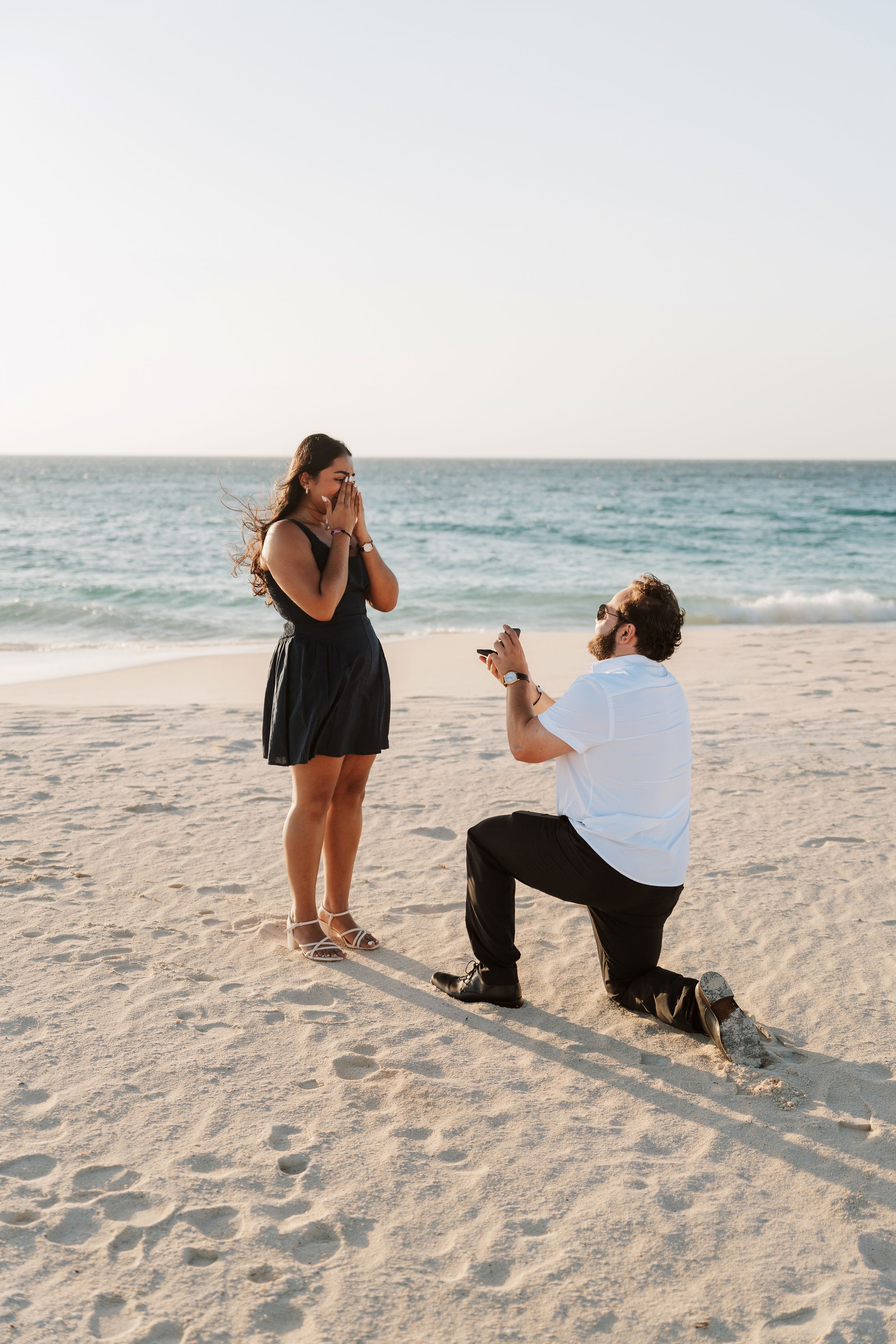 Man proposes on bended knee to woman on sandy beach at sunset, holding ring box as she covers her mouth in surprise.