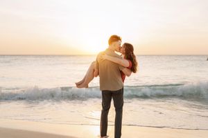 Romantic couple embracing at sunset on a beach, man lifting woman near ocean waves in a golden evening light.