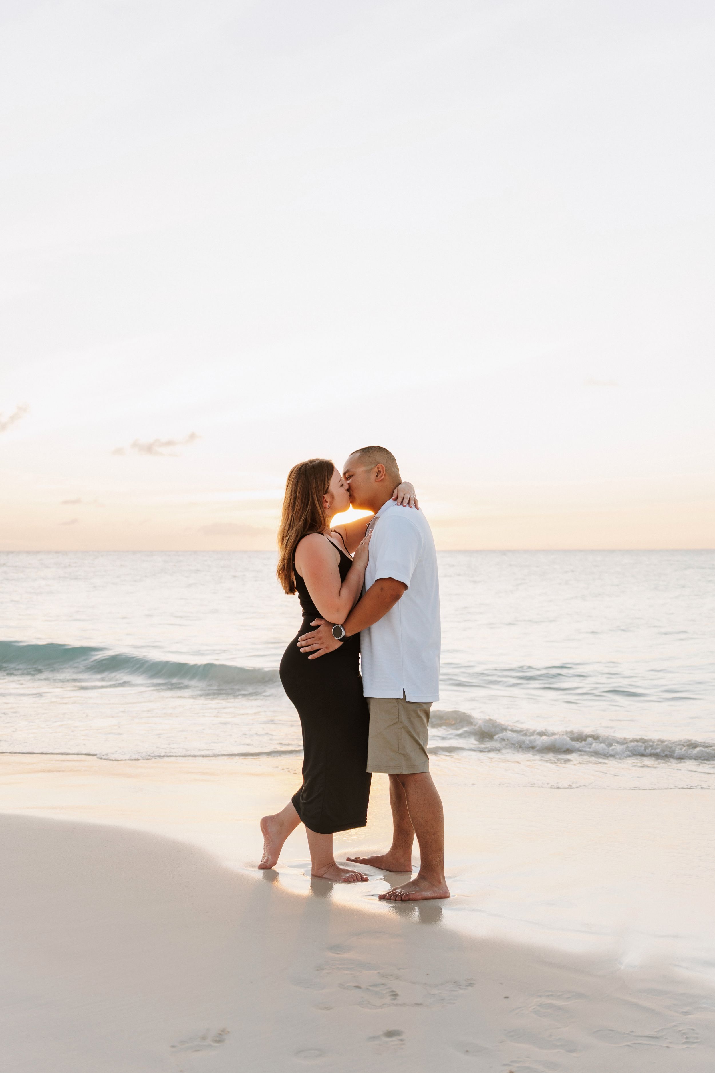 Couple kissing on a sandy beach at sunset, woman in black dress and man in white shirt and khaki shorts.
