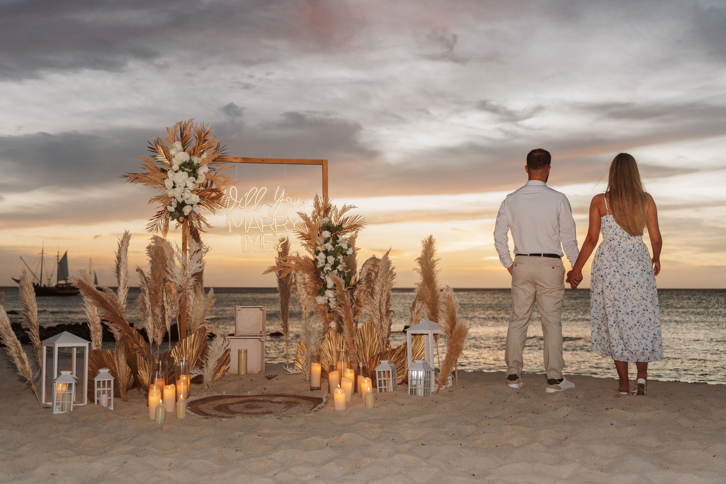 Couple holding hands at sunset beach proposal setup with pampas grass, lanterns, and floral arch on sandy shore.