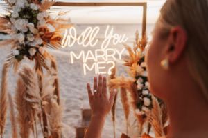 Woman touching a glowing neon "Will You Marry Me?" sign surrounded by floral arrangements at a beach proposal.