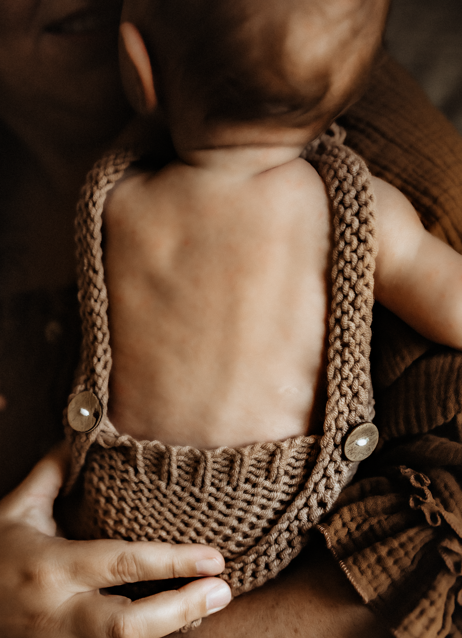 Baby wearing brown knit overalls with button straps, held by an adult, showing a close-up of the textured crochet romper.