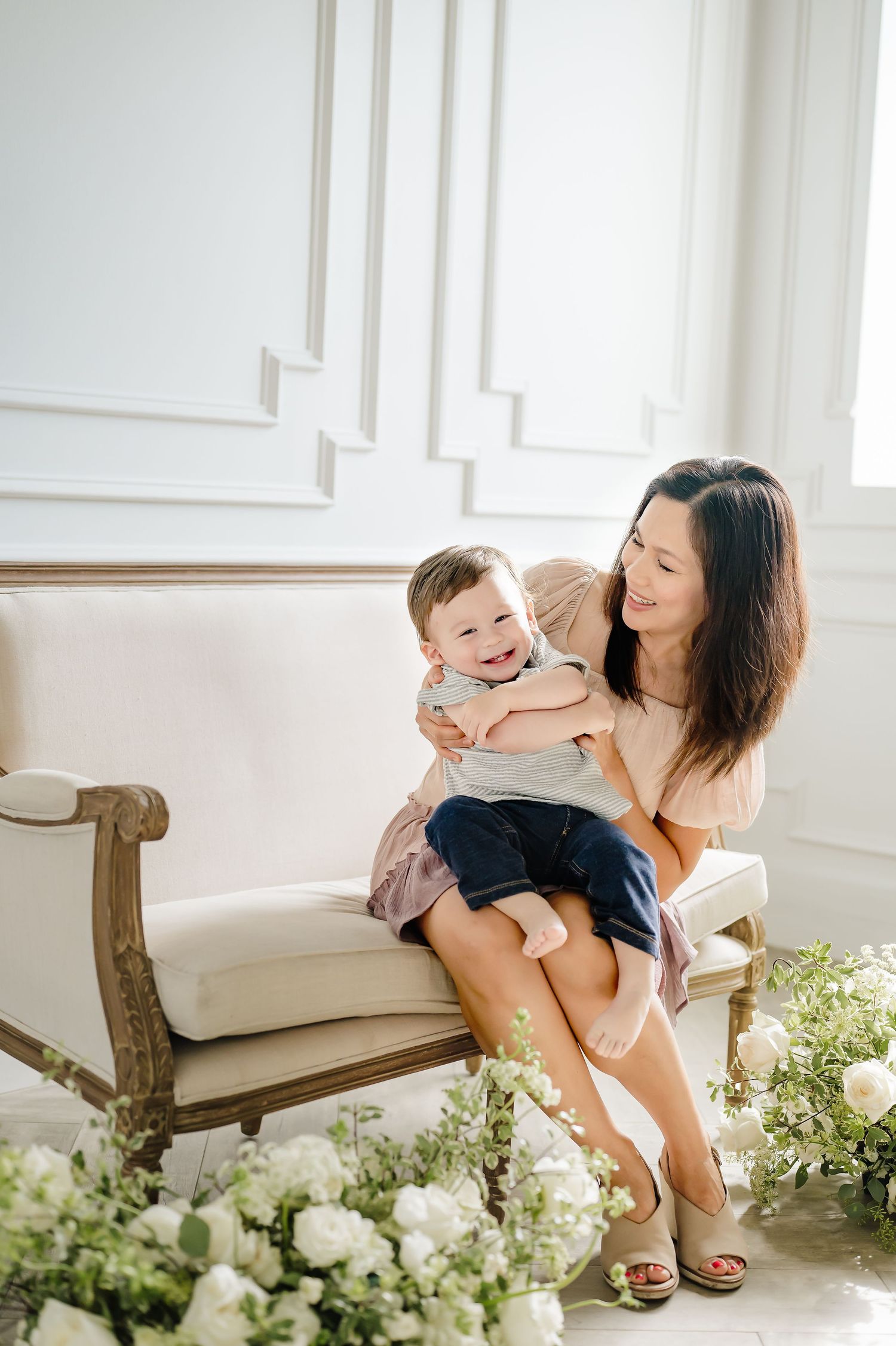 Playful mother-child interaction beside white floral arrangements in bright studio setting.