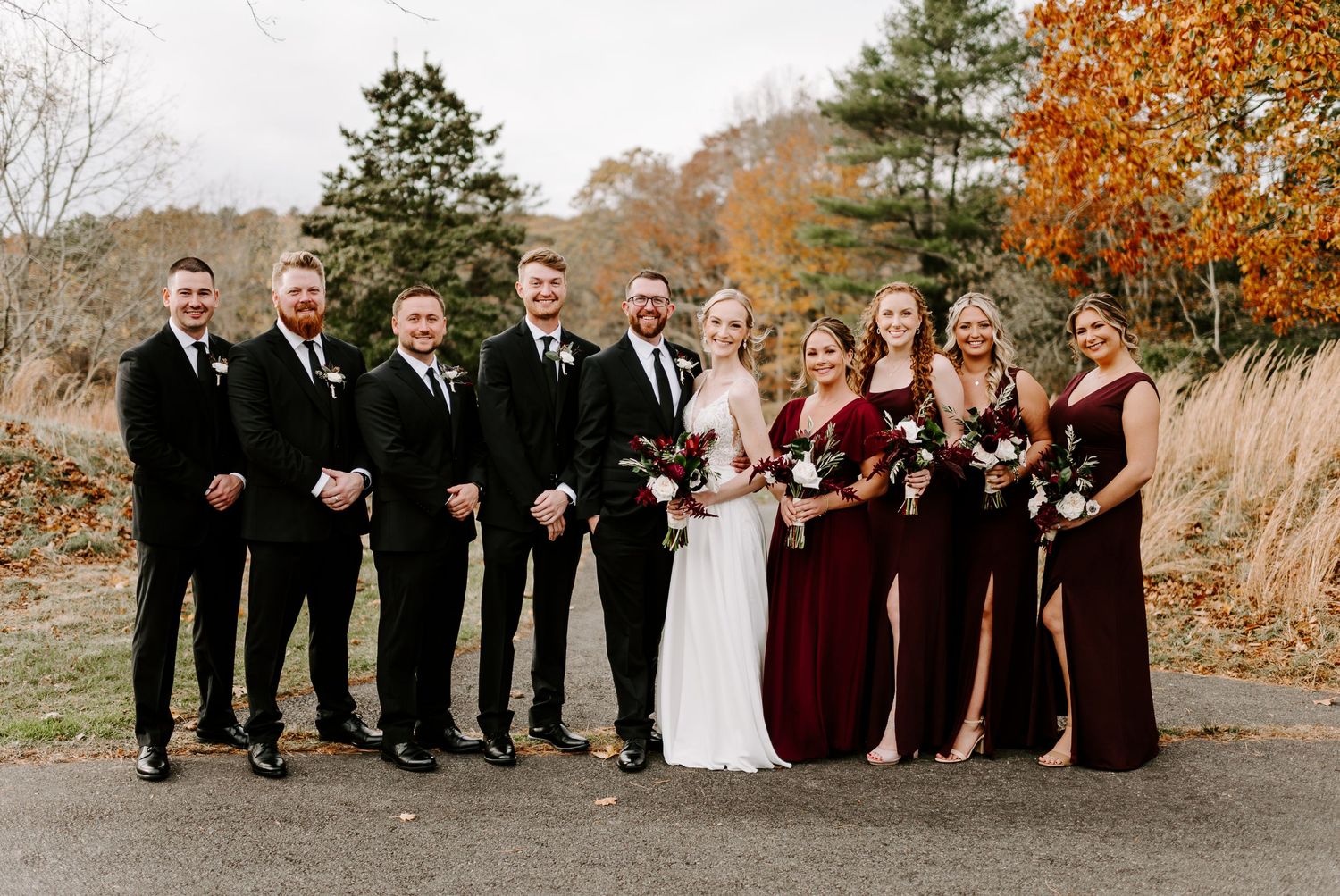 Wedding party members in formal attire line up for photo with autumn colors in background.