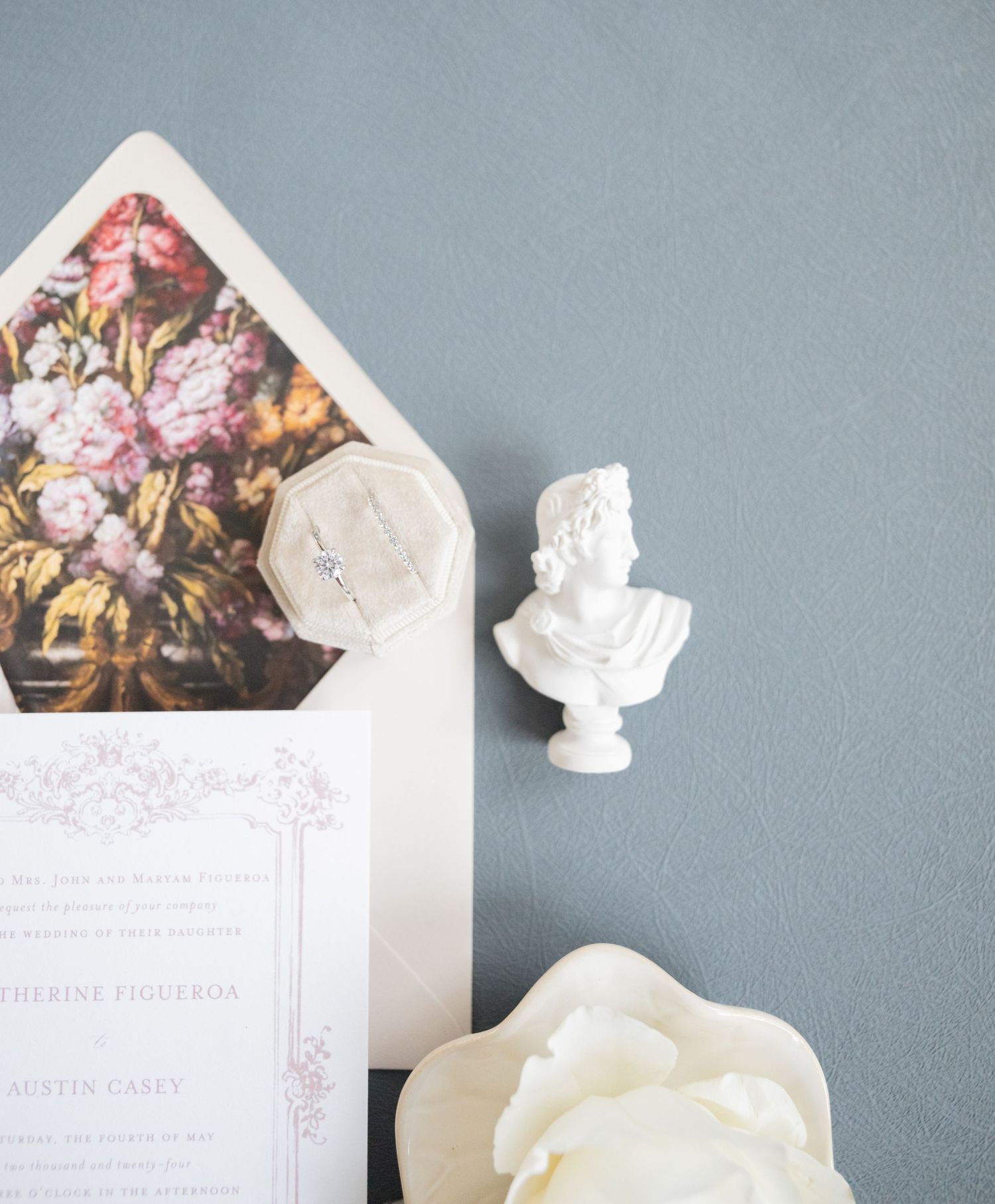 Floral lined envelope with wedding invitation and classical bust statue on grey background.