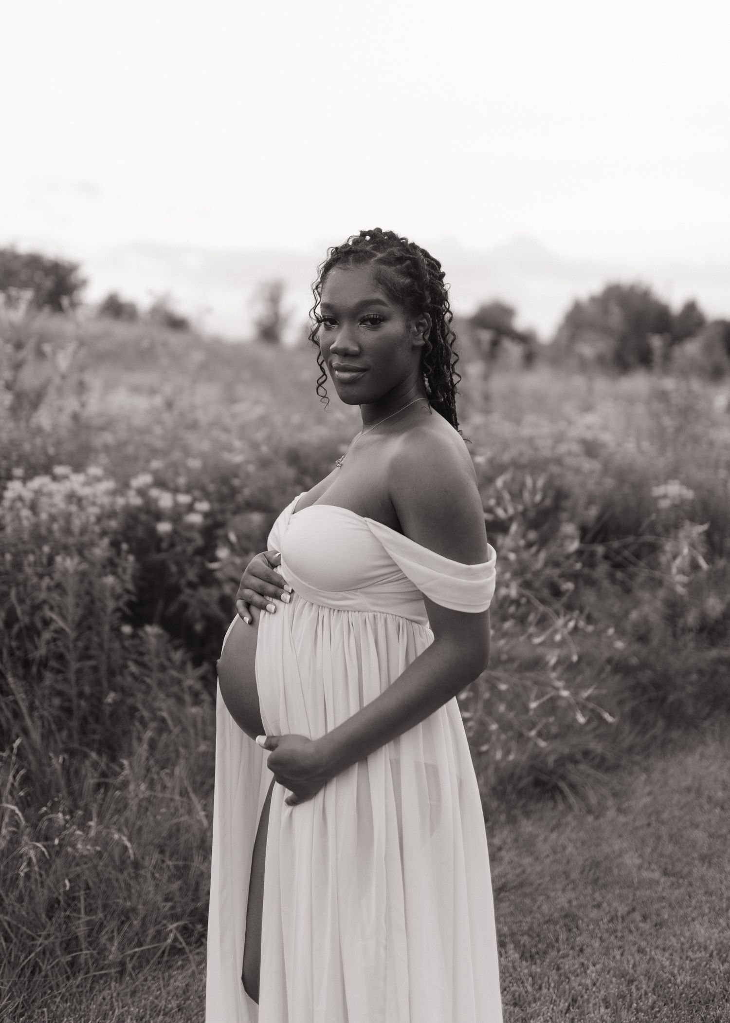 Black and white maternity photo of a pregnant woman in a white off-shoulder gown posing in a field in Northern California.