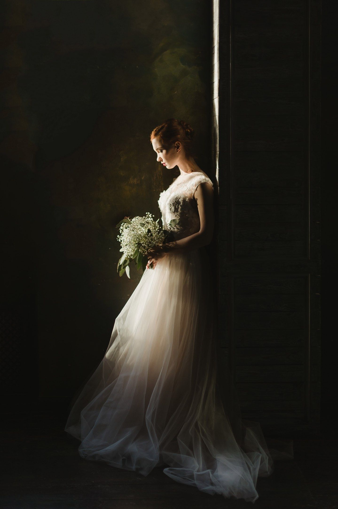 Elegant bride in a flowing white tulle gown holding a delicate bouquet, standing gracefully in soft dramatic lighting.