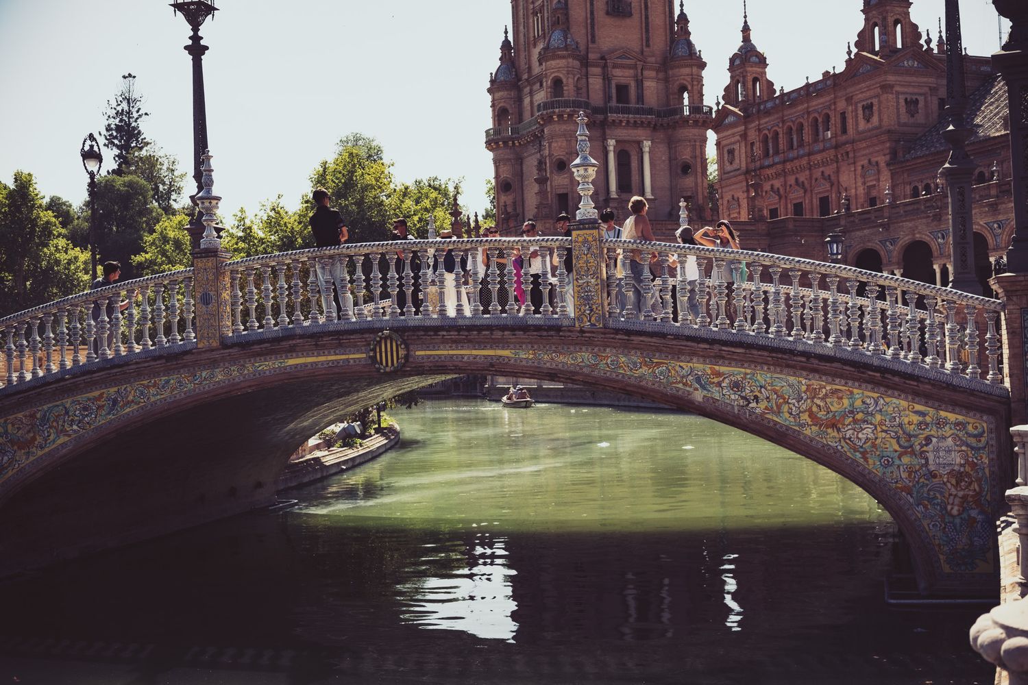 Ornate tiled bridge over green canal at Plaza de España, Seville, Spain, with tourists and grand baroque architecture.