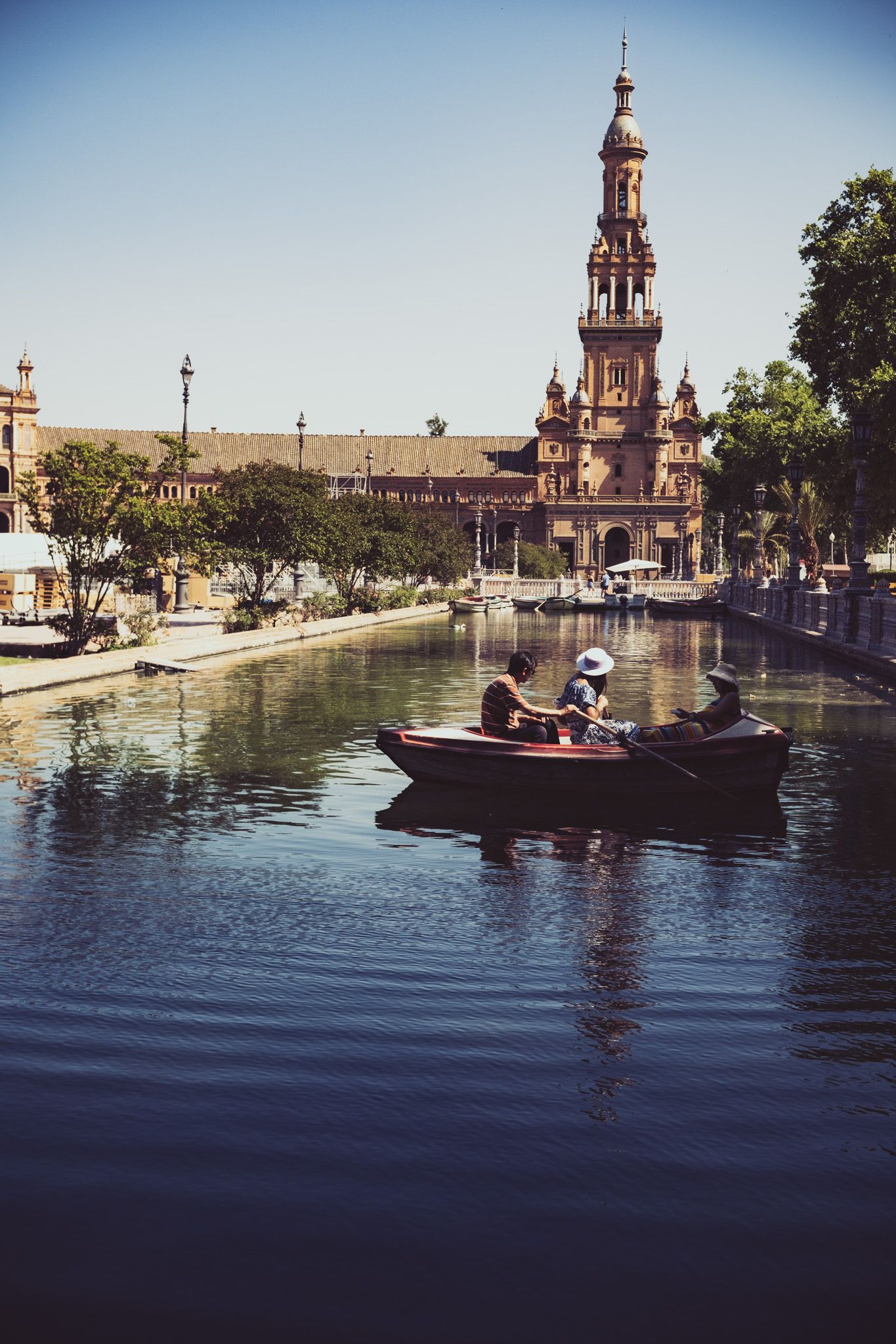 People rowing a wooden boat on the canal at Plaza de España with baroque tower in background, Seville.
