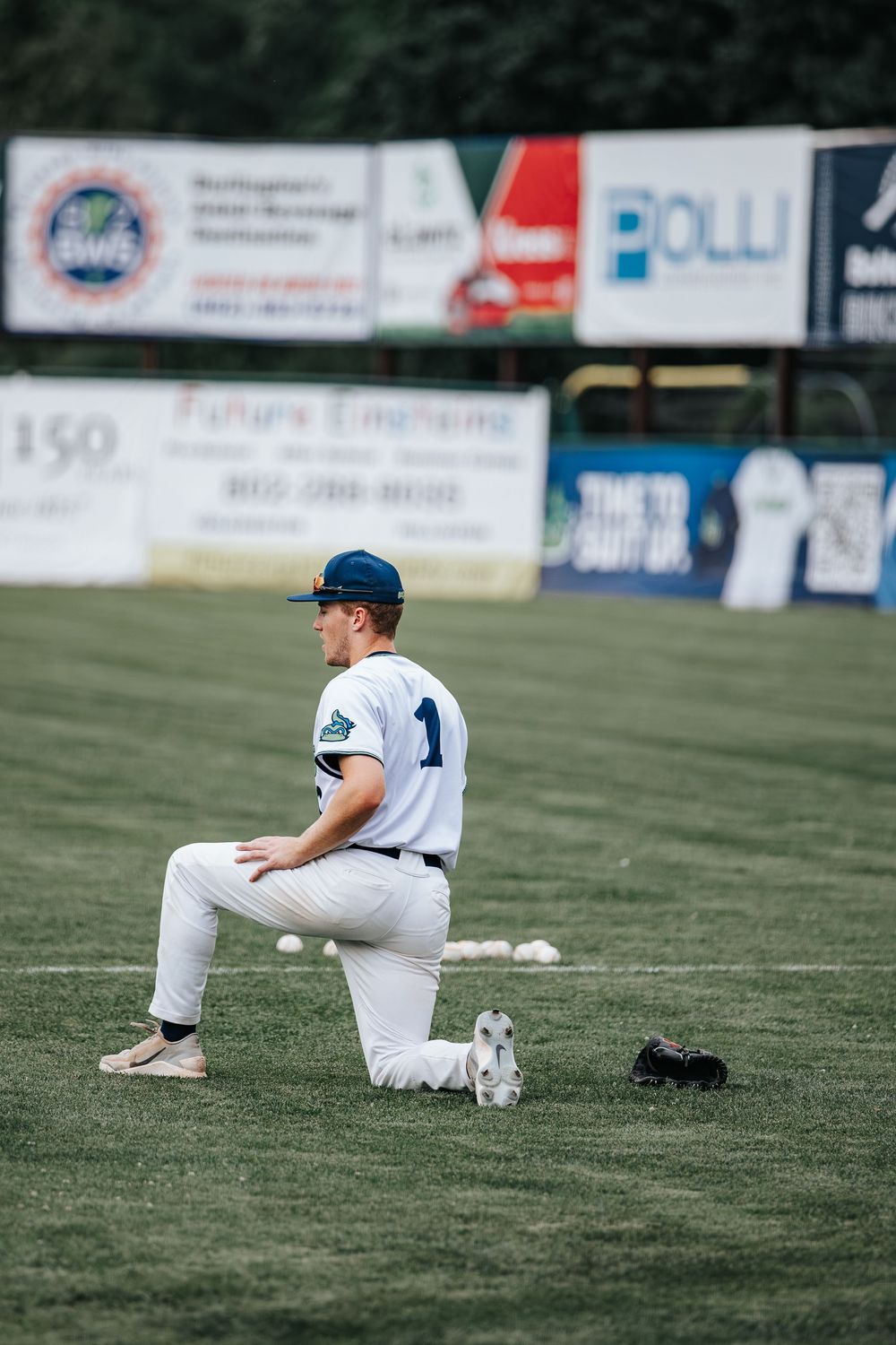 Baseball player in white uniform kneeling on green field during warm-ups with advertising banners in background.