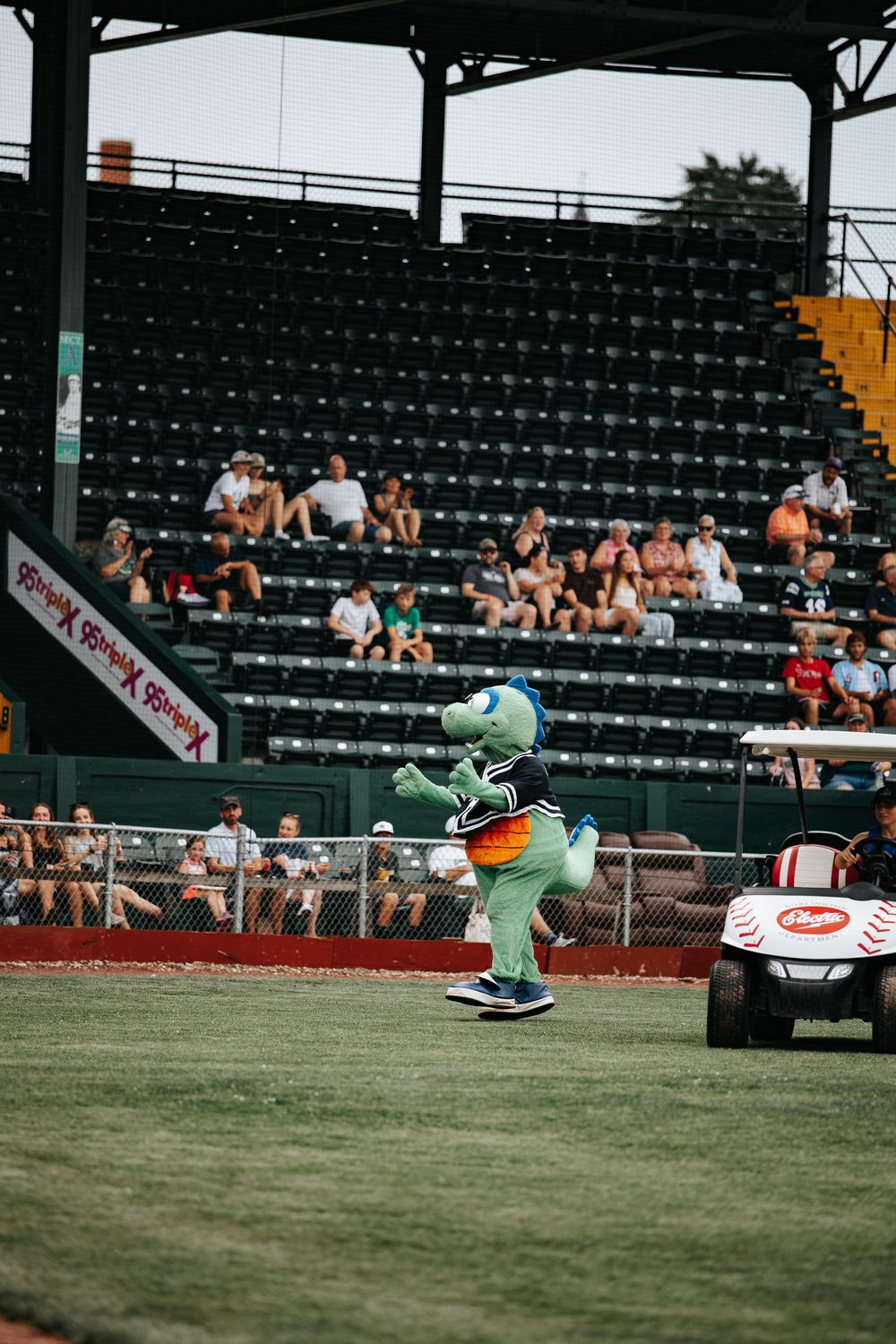 Green alien mascot dancing on baseball field at stadium with fans in bleachers during a minor league game.