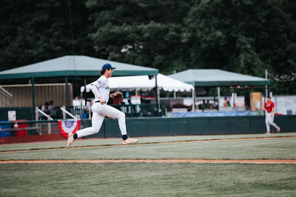 Baseball pitcher in white uniform mid-throw on green field with dugout and trees in background.