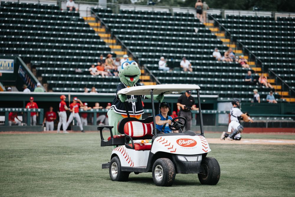 A baseball mascot in a golf cart rides on the field during a minor league baseball game at a green stadium.
