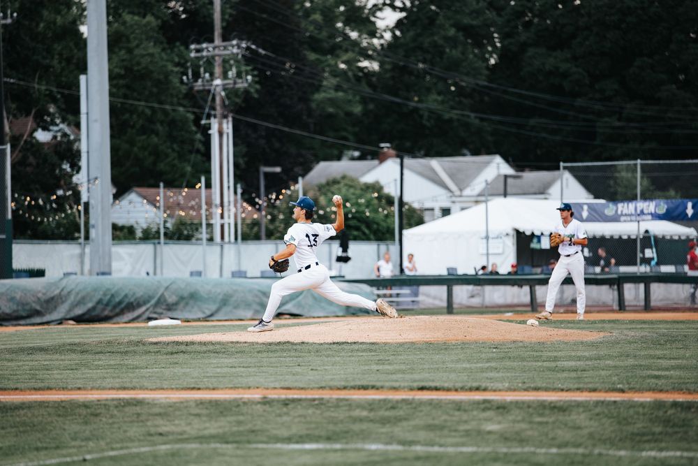 Baseball pitcher in mid-throw on green field with teammate and trees visible in background during game.