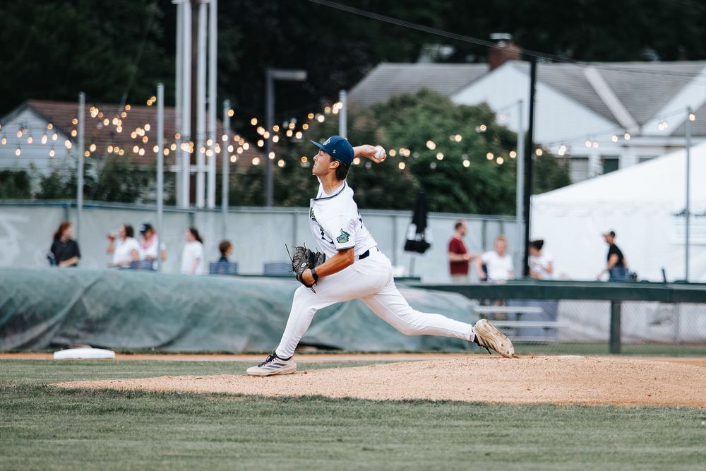 Baseball pitcher in white uniform mid-throw on mound at evening outdoor game with string lights in background.