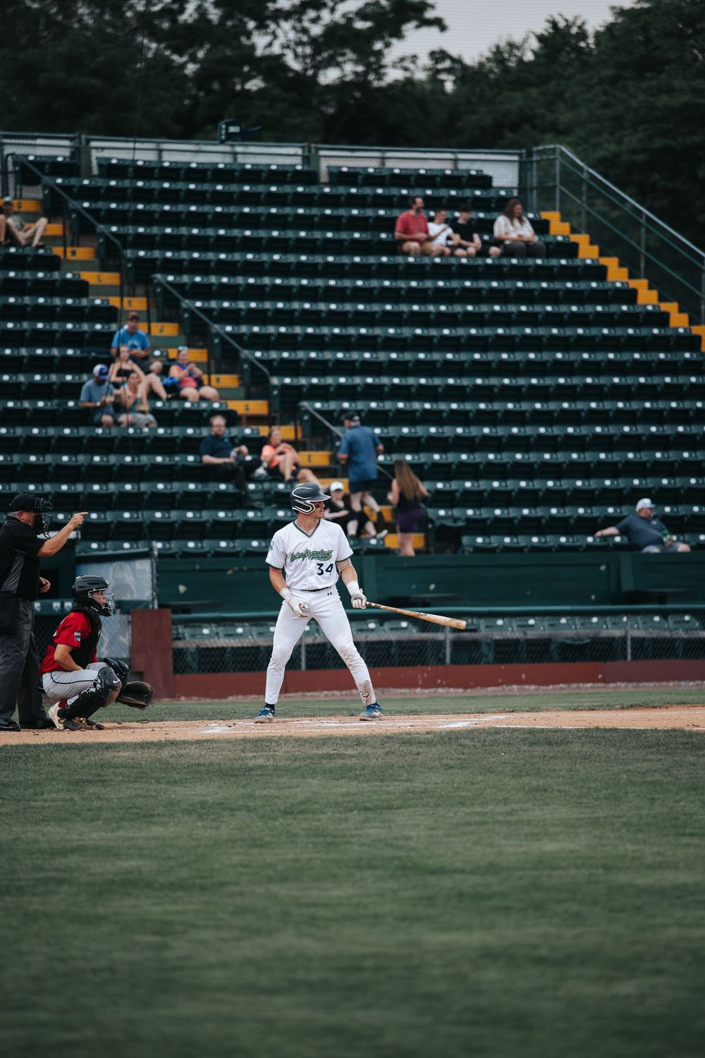 A baseball batter in white uniform stands ready at home plate during a game at a partially filled stadium.