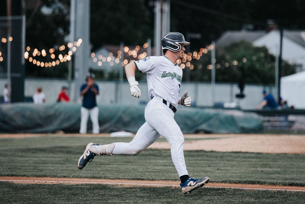 Baseball player in white uniform sprinting the bases during a night game with string lights in background.