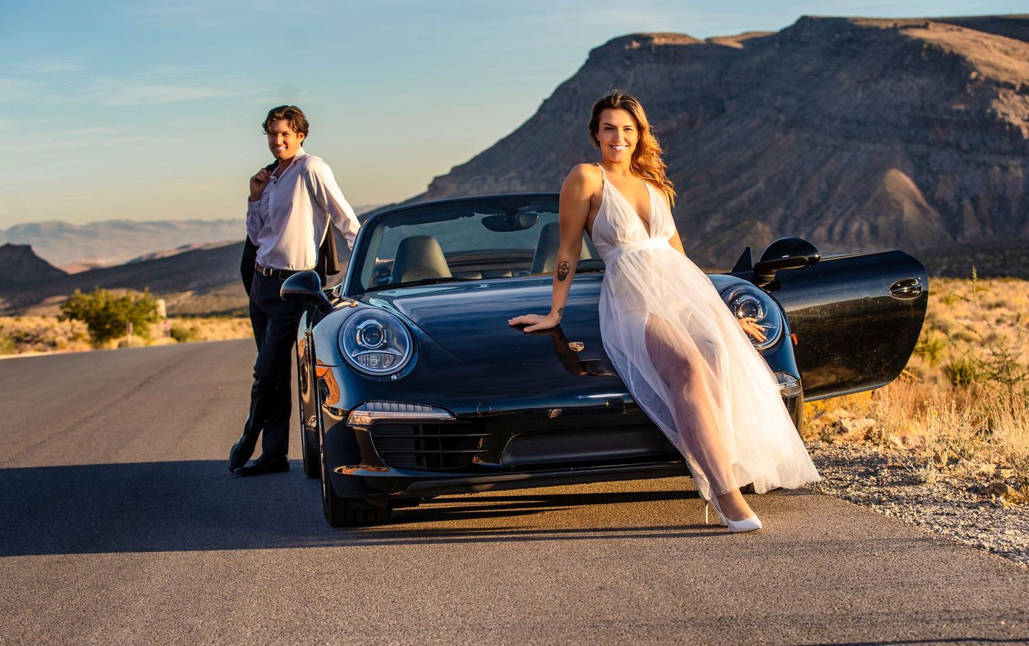 Couple in formal wear posing with a black Porsche 911 on a desert road with mountain backdrop.